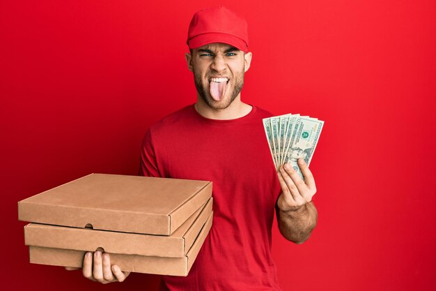 Giovane uomo caucasico che tiene cibo da asporto e 20 dollari che tirano fuori la lingua felice con un'espressione divertente