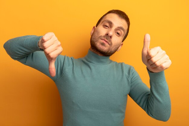 Giovane uomo caucasico dispiaciuto che guarda l'obbiettivo che mostra i pollici su e giù isolati su priorità bassa arancione