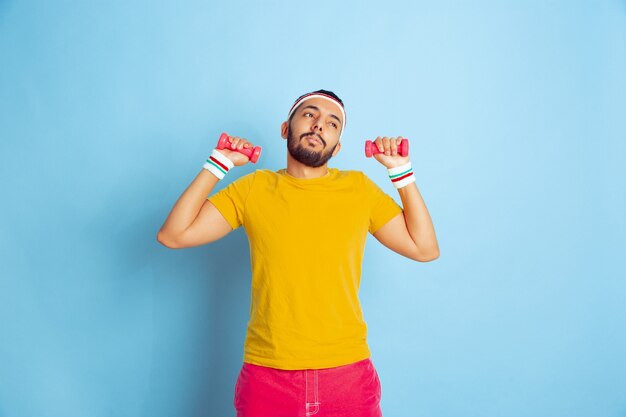 Giovane uomo caucasico in abiti luminosi, formazione su sfondo blu Concetto di sport, emozioni umane, espressione facciale, stile di vita sano, gioventù, vendite. Allenarsi con i pesi colorati. Copyspace.