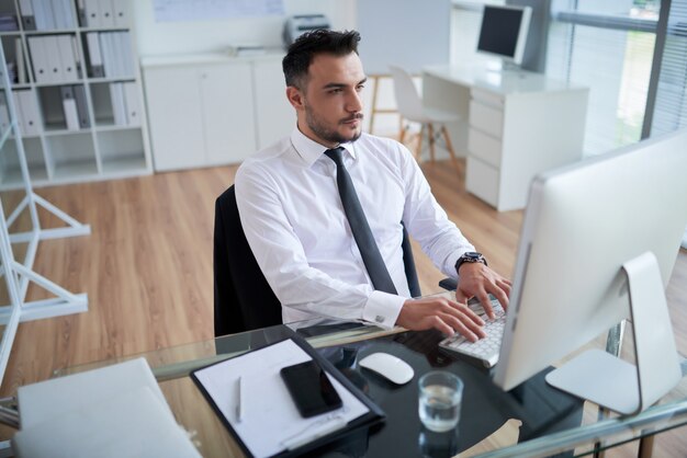 Giovane uomo caucasico in camicia formale e cravatta seduto in ufficio e lavorando sul computer