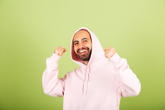 Giovane uomo caucasico in felpa con cappuccio rosa isolato, pugno di serraggio felice con successo di gesto del vincitore