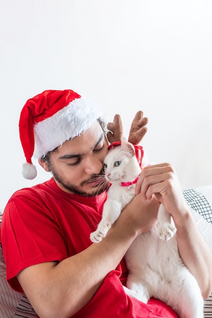 Giovane uomo caucasico in un cappello di Babbo Natale che gioca con il suo gatto bianco con le corna di alce