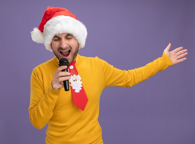 Giovane uomo caucasico indossando il cappello di Natale e cravatta tenendo il microfono cantando con gli occhi chiusi allungando la mano isolata su sfondo viola