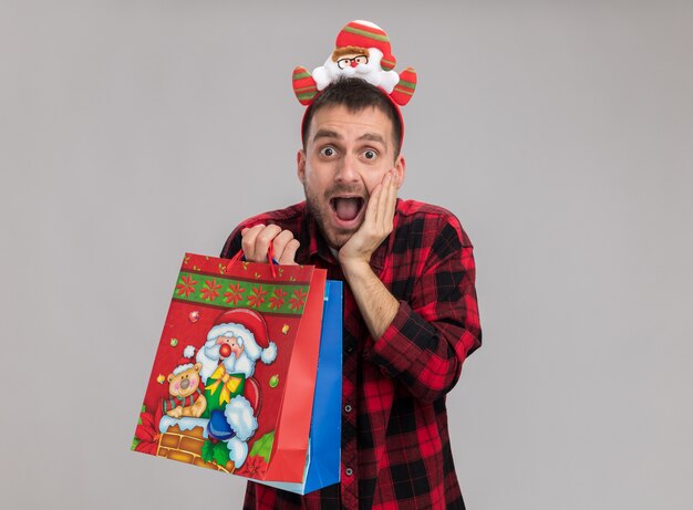 Giovane uomo caucasico scioccato che indossa la fascia di natale che tiene i sacchetti del regalo di Natale che esamina macchina fotografica che tiene la mano sul fronte isolata su fondo bianco con lo spazio della copia