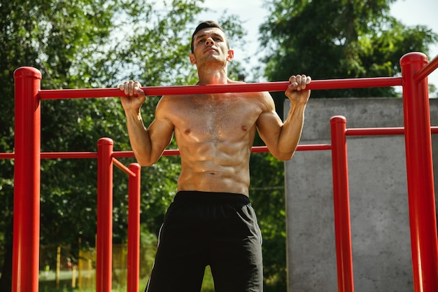 Giovane uomo caucasico senza camicia muscolare che fa pull-up sulla barra orizzontale al parco giochi nella soleggiata giornata estiva. Allenare la parte superiore del corpo all'aperto. Concetto di sport, allenamento, stile di vita sano, benessere.