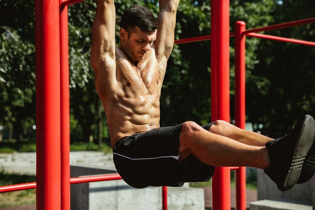 Giovane uomo caucasico senza camicia muscolare che fa pull-up sulla barra orizzontale al parco giochi nella soleggiata giornata estiva. Allenare la parte superiore del corpo all'aperto. Concetto di sport, allenamento, stile di vita sano, benessere.