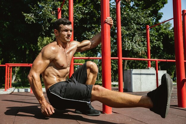 Giovane uomo caucasico senza camicia muscolare che fa squat vicino alla barra orizzontale al parco giochi nella soleggiata giornata estiva. Allenamento della parte inferiore del corpo all'aperto. Concetto di sport, allenamento, stile di vita sano, benessere.