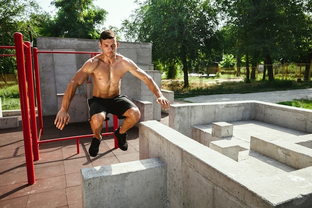 Giovane uomo caucasico senza camicia muscolare che salta sopra la barra orizzontale al parco giochi nella soleggiata giornata estiva. Allenare la parte superiore del corpo all'aperto. Concetto di sport, allenamento, stile di vita sano, benessere.