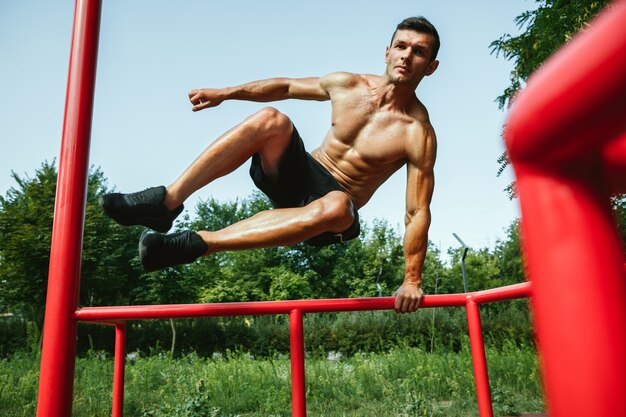 Giovane uomo caucasico senza camicia muscolare che salta sopra la barra orizzontale al parco giochi nella soleggiata giornata estiva