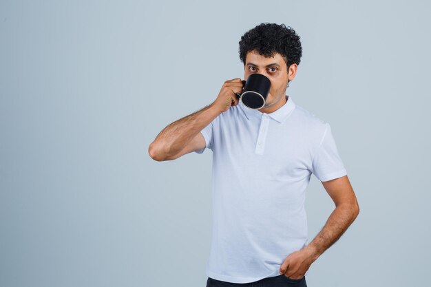 Giovane uomo che beve una tazza di tè mentre tiene la mano sulla vita in maglietta bianca e jeans e sembra serio. vista frontale.