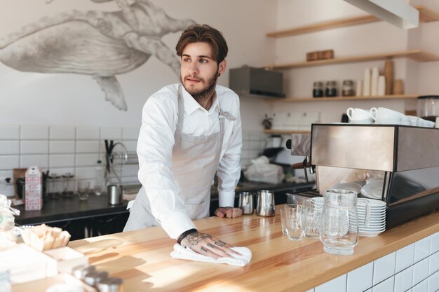 Giovane uomo che guarda premurosamente da parte e pulisce il bancone sul posto di lavoro nel ristorante Ritratto di barista in grembiule e camicia bianca in piedi al bancone della caffetteria