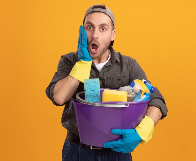 Giovane uomo che indossa abiti casual e berretto in guanti di gomma che tiene secchio con strumenti di pulizia guardando stupito e sorpreso in piedi sopra la parete arancione