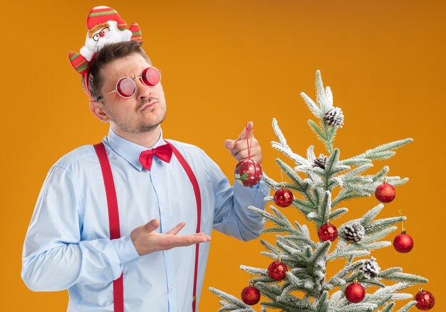 Giovane uomo che indossa bretelle farfallino in cappello della santa e occhiali rossi in piedi accanto all'albero di Natale sul muro arancione