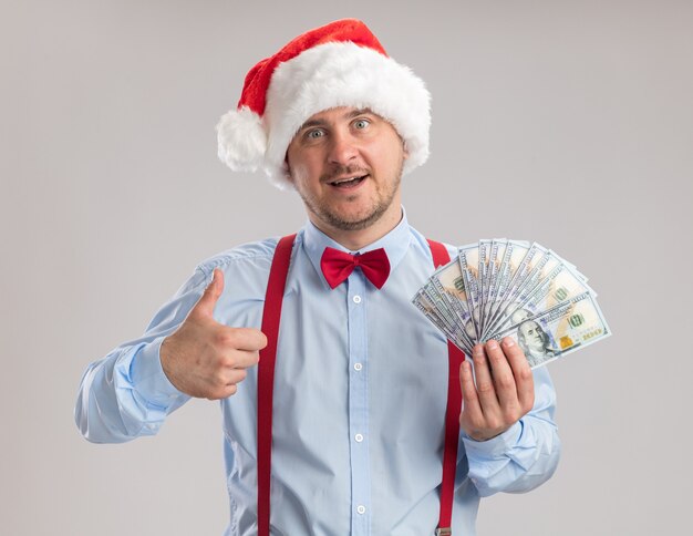 Giovane uomo che indossa bretelle farfallino in cappello di Babbo Natale che tiene contanti mostrando pollice in alto felice e sorpreso guardando la telecamera in piedi su sfondo bianco