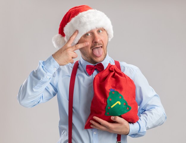Giovane uomo che indossa bretelle farfallino in cappello di Babbo Natale che tiene la borsa di Babbo Natale piena di regali guardando la fotocamera felice e allegro che mostra il segno v che attacca fuori la lingua in piedi su sfondo bianco