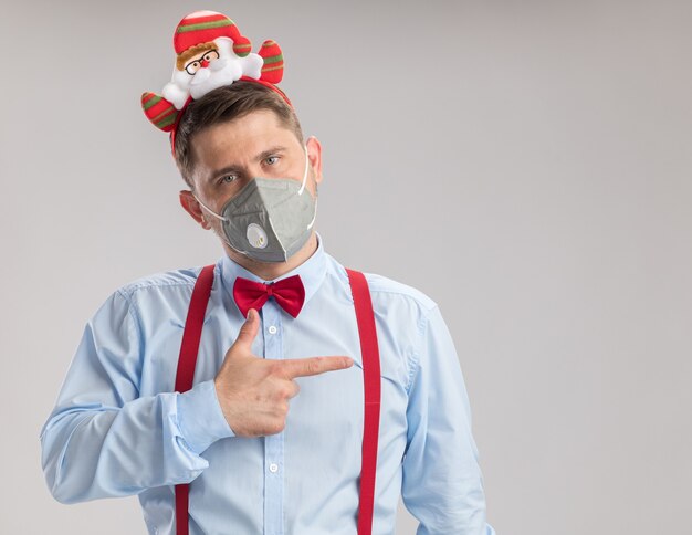 Giovane uomo che indossa bretelle farfallino in cerchio con santa che indossa una maschera facciale protettiva guardando la telecamera che punta con il dito indice sul lato in piedi su sfondo bianco