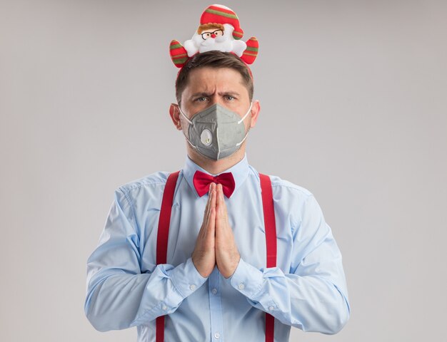 Giovane uomo che indossa bretelle farfallino in cerchio con santa che indossa una maschera facciale protettiva guardando la telecamera con un viso serio che tiene le mani insieme come pregare in piedi su sfondo bianco