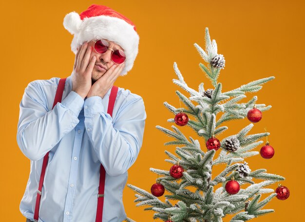 Giovane uomo che indossa bretelle farfallino in santa hat e occhiali rossi in piedi accanto all'albero di natale guardando la telecamera confuso e molto ansioso su sfondo arancione