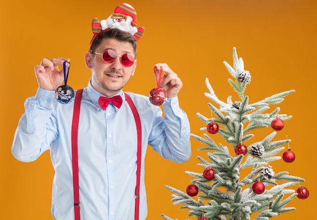 Giovane uomo che indossa bretelle farfallino nel cerchio con Babbo Natale e occhiali rossi in piedi accanto all'albero di natale cercando confuso tenendo i giocattoli per albero su sfondo arancione