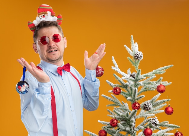 Giovane uomo che indossa bretelle farfallino nel cerchio con Babbo Natale e occhiali rossi in piedi accanto all'albero di natale cercando confuso tenendo i giocattoli per albero su sfondo arancione