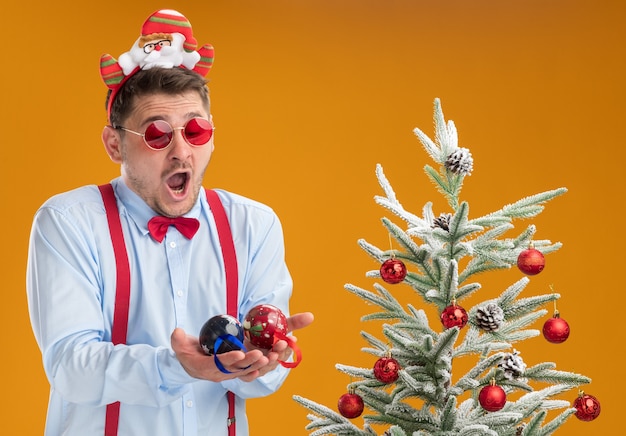 Giovane uomo che indossa bretelle farfallino nel cerchio con Babbo Natale e occhiali rossi in piedi accanto all'albero di Natale che sembra confuso e stupito tenendo i giocattoli per albero sopra la parete arancione