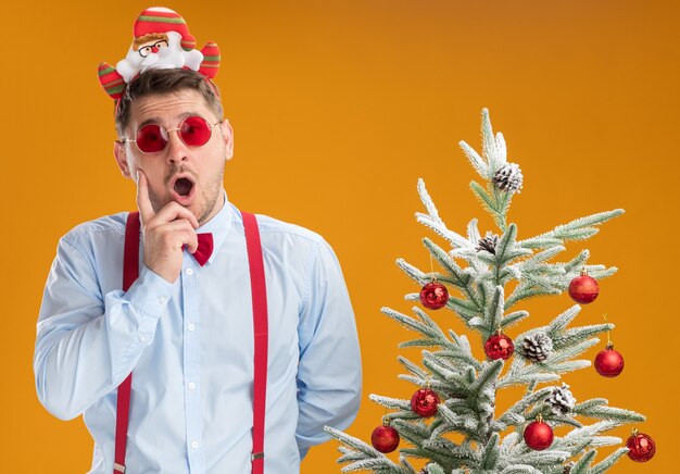 Giovane uomo che indossa bretelle farfallino nel cerchio con Babbo Natale e occhiali rossi in piedi accanto all'albero di natale guardando la telecamera stupito e sorpreso su sfondo arancione