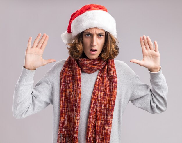 Giovane uomo che indossa il natale santa hat con calda sciarpa intorno al collo guardando la telecamera con grave accigliata faccia con le braccia alzate in piedi su sfondo bianco