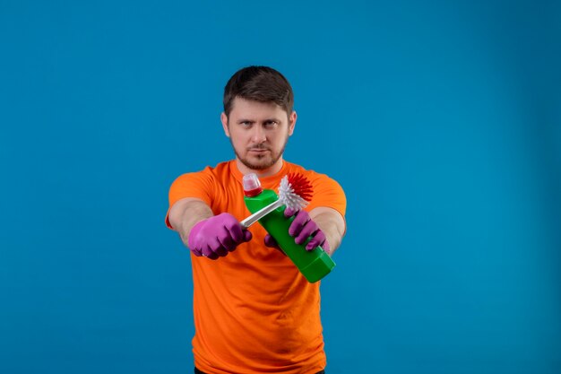 Giovane uomo che indossa la maglietta arancione e guanti di gomma che tengono i prodotti per la pulizia e la spazzola