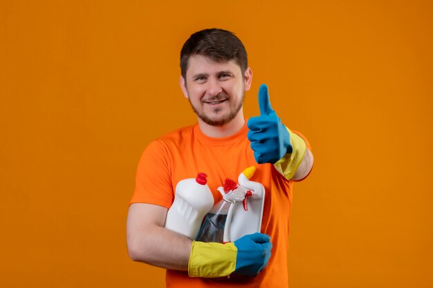 Giovane uomo che indossa la maglietta arancione e guanti di gomma tenendo i prodotti per la pulizia sorridendo allegramente positivo e felice guardando la telecamera che mostra i pollici in su pronto per pulire il concetto su sfondo arancione