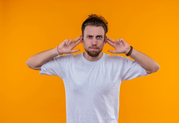 giovane uomo che indossa la maglietta bianca mise le dita sulle orecchie sul muro arancione isolato