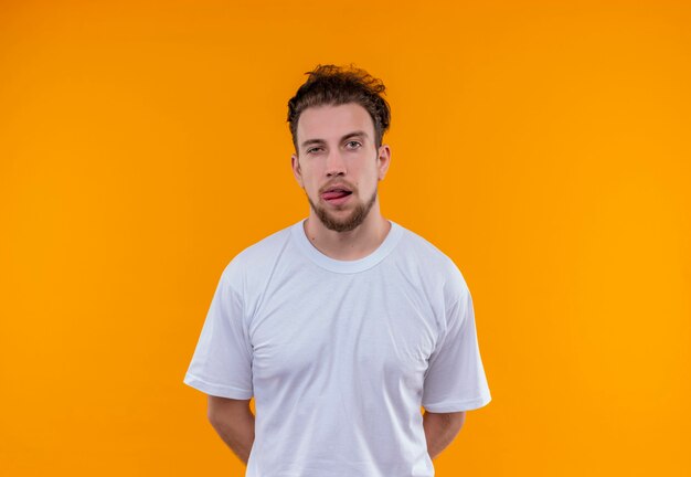 giovane uomo che indossa la maglietta bianca mise le mani sulla schiena e mostrando la lingua sul muro arancione isolato