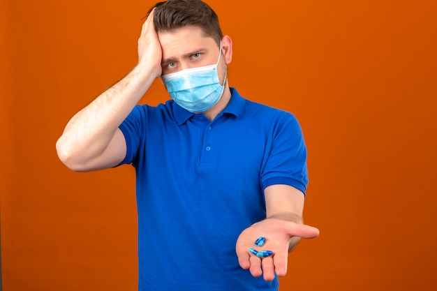 Giovane uomo che indossa la polo blu in maschera protettiva medica guardando malato e malato in piedi con la mano sulla testa che soffrono di mal di testa con bucce aperte a mano su muro arancione isolato