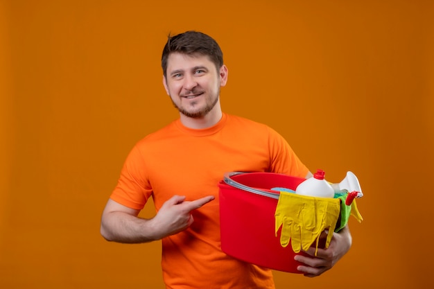 Giovane uomo che indossa la t-shirt arancione tenendo la benna con strumenti di pulizia puntando con il dito ad esso sorridente guardando la telecamera positivo e felice pronto per la pulizia in piedi su sfondo arancione
