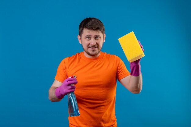 Giovane uomo che indossa maglietta arancione e guanti di gomma che tengono spray per la pulizia e spugna
