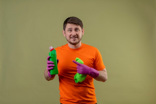 Giovane uomo che indossa maglietta arancione e guanti di gomma che tiene spray per la pulizia e tappeto che punta con il dito per imbottigliare con spray sorridente allegramente positivo e felice guardando la fotocamera su sfondo verde