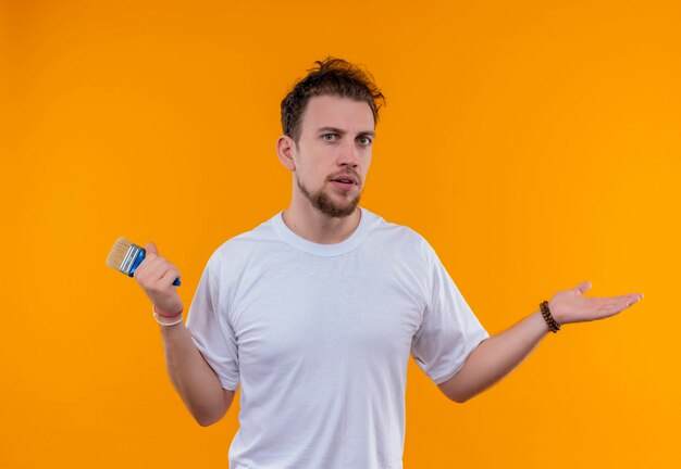 giovane uomo che indossa t-shirt bianca tenendo il pennello e mostrando quale gesto sulla parete arancione isolata