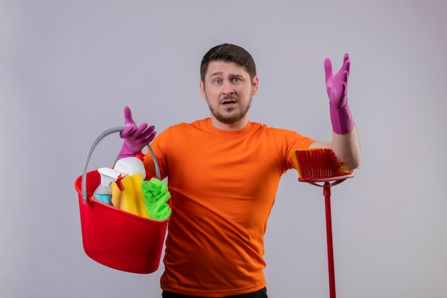 Giovane uomo che indossa una maglietta arancione e guanti di gomma tenendo la benna con strumenti di pulizia e mop scontento con l'espressione confusa sul viso in piedi sopra il muro bianco