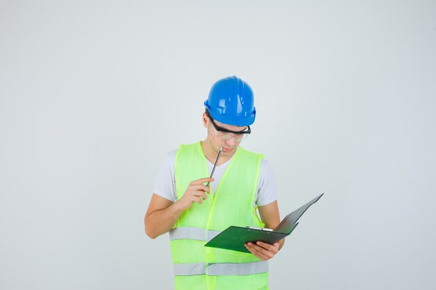 Giovane uomo che legge le note negli appunti, mettendo la penna vicino alla bocca in uniforme da costruzione e guardando a fuoco, vista frontale.