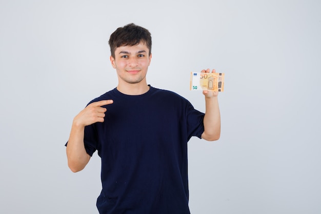 Giovane uomo che punta alla banconota in euro in maglietta nera e guardando fiducioso, vista frontale.