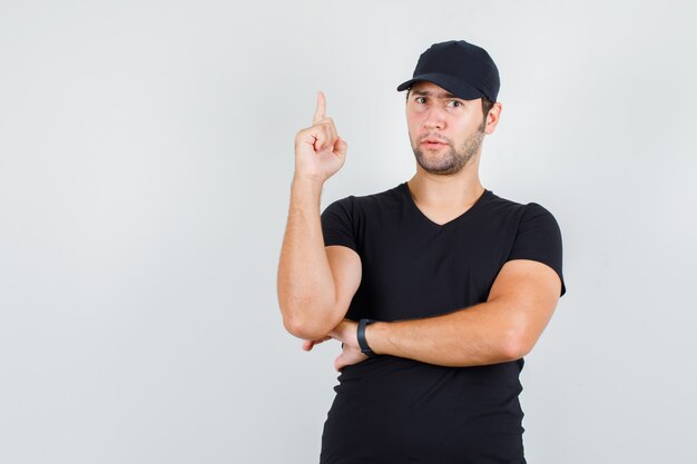 Giovane uomo che punta il dito verso l'alto in maglietta nera, berretto e guardando curioso.