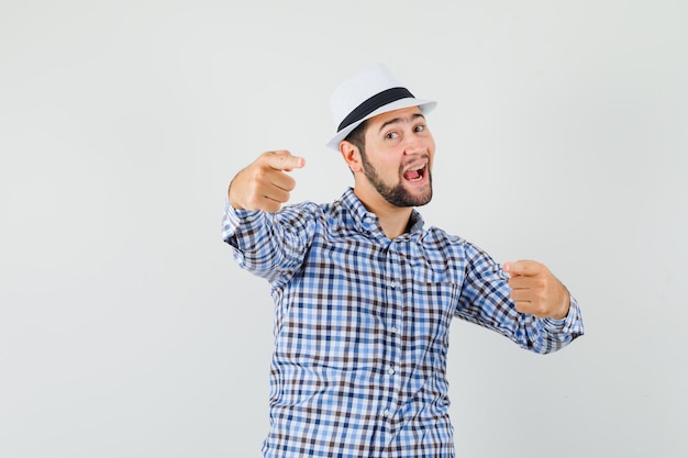 Giovane uomo che punta le dita alla fotocamera in camicia a quadri, cappello e guardando felice, vista frontale.