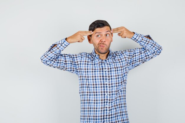 Giovane uomo che punta le mani e le dita alla testa in camicia a quadri e guardando pensieroso.