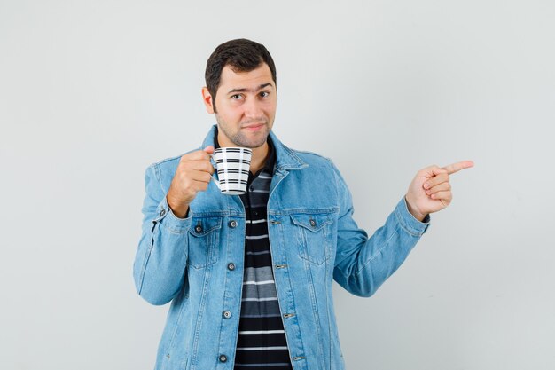 Giovane uomo che punta lontano, tenendo la tazza di bevanda in t-shirt, giacca e guardando fiducioso, vista frontale.