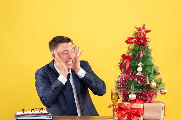 giovane uomo chiudendo le orecchie con le dita seduto al tavolo vicino all'albero di Natale e regali su giallo