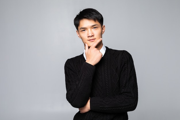 Giovane uomo cinese asiatico con le mani sul mento che controlla parete bianca isolata