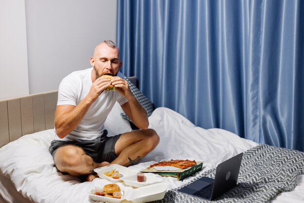 Giovane uomo con fast food a casa in camera da letto sul letto