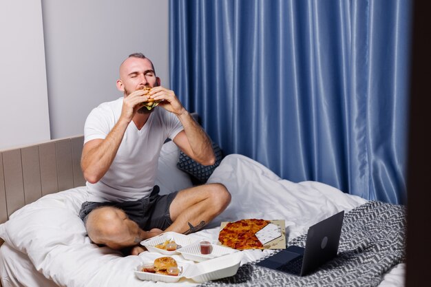 Giovane uomo con fast food a casa in camera da letto sul letto