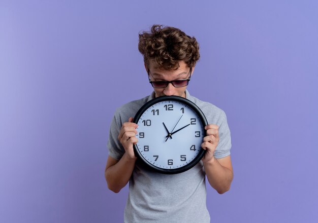 Giovane uomo con gli occhiali neri che indossa la maglietta polo grigia che tiene orologio da parete che sembra preoccupato in piedi sopra la parete blu