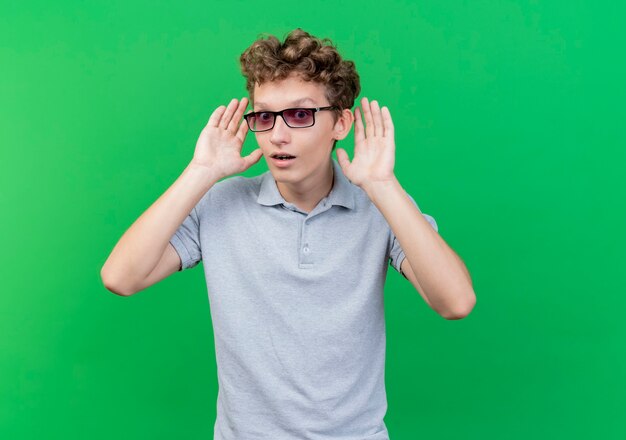 Giovane uomo con gli occhiali neri che indossa la maglietta polo grigia sorpreso e felice con le braccia vicino alle orecchie in piedi sul muro verde