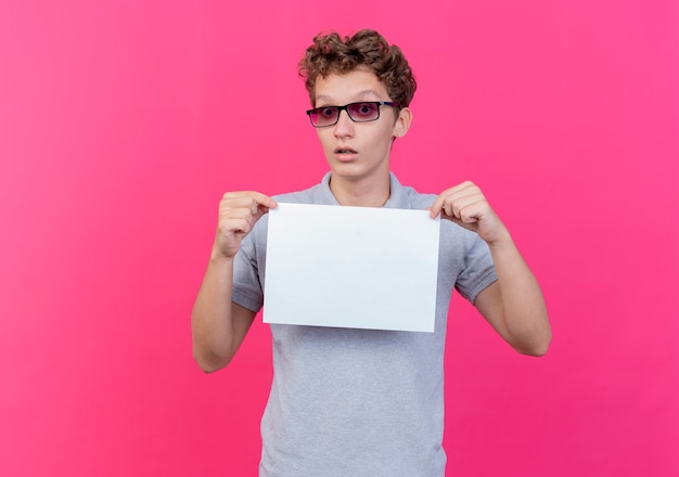 Giovane uomo con gli occhiali neri che indossa una polo grigia che mostra un foglio di carta bianco sorpreso in piedi sul muro rosa